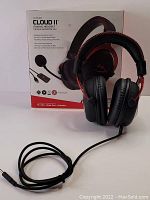 Photo showing HyperX Cloud II headset positioned in front of its branded retail box. The headset is black with red accents, featuring cushioned over-ear cups and an attached cable ending in a 3.5mm jack.