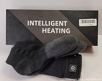 Photo of one pair of black and grey Intelligent Heating brand rechargeable heated socks displayed on top of the original packaging box showing product and branding clearly.