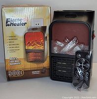 Photo shows the flame heater unit alongside its original packaging box and a plastic-wrapped remote control in front, depicting the heater's appearance and features.