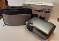 Photo of the Jeemak P100 projector alongside its carrying case and original box on a white surface.