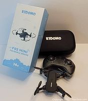 Photo shows Kidomo F02 Mini foldable drone, its remote controller, carrying case and original box placed on surface.