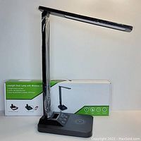 Photo of black foldable desk lamp with touch control panel and wireless charging base, alongside original packaging box showing product image and key features.