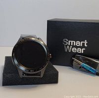 Black metal smart watch displayed on a foam holder with round digital screen and numerical bezel. Next to it are a charging cable and a black box with Smart Wear branding.
