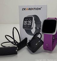 Photo showing Zkoreation smart watch with purple and black silicone bands, two charging docks with USB cables, and the original product box with brand and watch image.