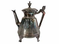 Front and side perspective of the silver teapot showing Egyptian Revival design, spout, handle, decorative finial lid, and triple leg base.