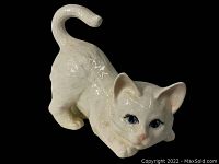 Side angle of a white glossy ceramic cat figurine with detailed fur texture and blue eyes in a crouching pose.