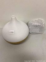 Photo showing white ribbed aromatherapy diffuser with narrow top nozzle and wooden base beside bubble-wrapped power adapter.