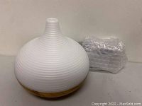 Photo of the white and wood aromatherapy diffuser with its black power adapter wrapped in bubble wrap on a white surface.