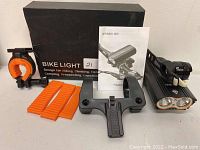 Black dual-beam bike light with mounting bracket, orange rubber straps, phone mount, and box with user manual.