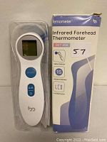 White infrared forehead thermometer with LCD screen and blue buttons displayed alongside original product box labeled DET-306.