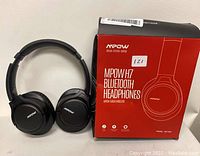 Pair of black MPOW H7 Bluetooth wireless headphones positioned next to their original red and black retail packaging box showing the brand and model information.