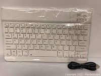 Photo of a white ultra slim Bluetooth keyboard measuring 10 inches, showing the full QWERTY key layout and function keys. A black USB cable is placed alongside for charging or wired connection.