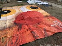 Large red and orange printed tarp featuring a stylized face and text 'LOVE AT THE BOTTOM OF THE SEA' laid out flat on ground.
