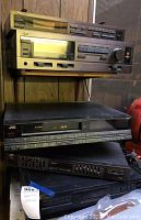 Four stacked JVC audio and video components, including stereo receiver, optical disc player, and graphic equalizer, with wood panel wall backdrop.