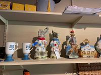 Shelf with diverse Beam bourbon whiskey collectible decanters including dog-shaped, historical figurines, and commemorative themed decanters alongside two mugs with blue bases.