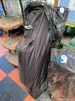 Black carrying bag standing upright, showing handle and exterior pocket, containing folded canopy tent and frame components.