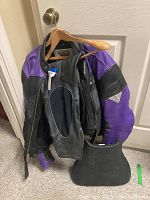 Firstgear black and purple leather motorcycle jacket with mesh vents, hanging on a door hook, showing condition and color details alongside a glimpse of black leather pants under the jacket.