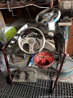 Full view of three video game steering wheels on a metal stand with pedals below and a red metal bowl on the rack shelf.