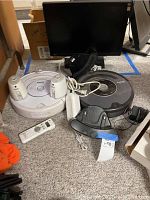 Photo showing two iRobot Roomba robot vacuum cleaners on carpet, one white and one dark gray, with associated charging bases, power cords, and a remote control. Background includes a monitor and some boxes.