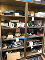 Wooden shelving unit with multiple shelves holding a variety of electronic gadgets and accessories including the boxed Veho camcorder, cameras, cables, a keyboard, mouse, and more.