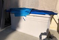 White plastic outdoor storage bin containing swimming pool gear including float mats and pool float in box, partially covered by fabric