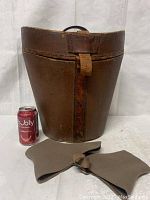 Front view of brown leather antique hat box with handle and leather strap closure, alongside brown wool spats laid flat, shown with soda can for size reference.