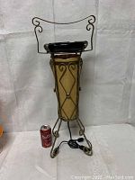 Full view of the tall electric ashtray stand with a black ashtray on top, beige fabric panel insert, and twisted metal scroll frame. A cherry Bubly soda-can shown for scale.