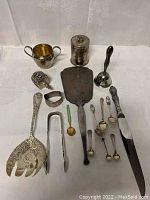 Overview photo of lot laid out on table showing all items together including spoons, tongs, serving pieces, candlestick, container, cup.