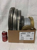 Front view of multiple vintage metal film canisters inside a shipping box beside a can of soda for scale, showing varying sizes and colors of lids.