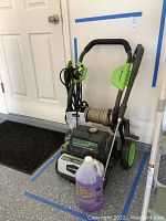 Full view of Greenworks electric pressure washer unit with hose rolled on reel, spray gun attached with power cord wrapped, placed next to a purple bottle of soap.