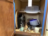 Cupboard shelf with assortment of metal baking pans, pizza stone, wooden rolling pin, glass and plastic measuring cups, metal measuring spoons, and Tupperware cake carrier on top shelf.