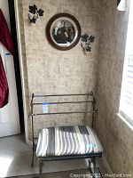 Photo of bathroom towel stand with padded stool in front and wall decor including round framed plate and two metal votive sconces.