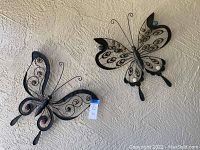 Two metal butterfly sculptures mounted on a textured wall, showing size and detailed wing patterns with colored circular accents