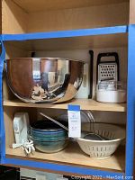 Shelves holding large stainless steel mixing bowl, hand mixer, plastic cutting boards, black grater with white base, and small enameled mixing bowls with metal colander.