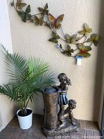 Full view of fountain with boy and girl statue, live palm plant in white pot, and butterfly metal wall art on beige wall.