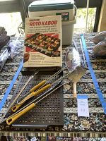 Image showing all grill accessories laid on patterned cloth, including Roto-Kabob box, skewers, spatula, tongs, and Igloo cooler in the background.