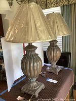 Pair of bronze urn-shaped table lamps with beige, plastic-covered fabric shades, shown on quilted surface with white divider backdrop.