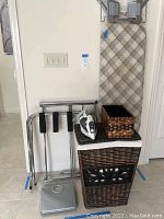 Photo showing ironing board leaning against wall with woven laundry hamper, metal drying rack, luggage valet stand, and bathroom scale.