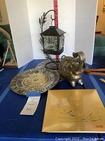 Group photo showing all decorative metal tone pieces including the lantern on curved stand, elephant figurine, metal inlaid plate, and painted wood plate