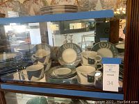 Full view of Mikasa Intaglio stoneware dishes arranged inside a glass curio cabinet, showing cups, plates, and small bowls.