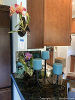 Two 12-inch artificial orchid wall hangings mounted on driftwood plaques standing upright next to a black metal candelabra with three turquoise candles. Decorative square glass plate with floral design visible behind.