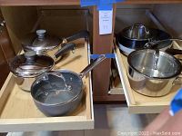 Pans and skillets stored in wooden kitchen cabinet drawers, showing handles, lids, and pot sizes.