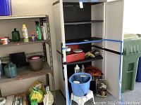 Plastic storage cabinet with five black shelves inside. White ribbed doors with black handles are open showing interior shelves and storage space. Cabinet stands next to metal shelving and garage tools visible in background.