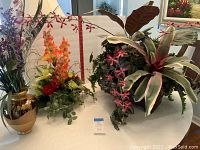 View of four silk floral arrangements on white table including tall colorful flowers in gold and clear vases and tropical themed silk plants.