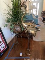 Full view showing tall gold-colored metal bird sculpture next to silk plant in geometric-patterned leather look pot on metal stand.
