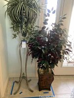 Full view of two silk plant arrangements showing the silver tone planter on a tripod stand and the bronze tone square planter with foliage