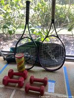 Two tennis rackets upright with cases behind, four pairs of red hand weights, and can of Wilson tennis balls on floor with outdoor background