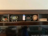Shelved view showing three black tole mini trays with colorful floral designs, a brass inlaid round plate, and a rectangular brass inlaid box.
