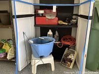 Photo shows plastic step stool with blue bucket on top. Extension cords visible on bottom shelf, nails in a white container on floor, and cleaning liquids.