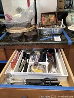 Photo of drawer with assorted cooking utensils organized in a white plastic tray including whisk, can opener, tongs, garlic crusher